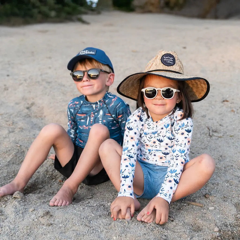 Two young children wearing a long-sleeve rash guard and hat, playing in full sunlight at the beach