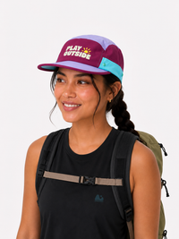 Woman wearing a colorful pink and purple athletic hat with 'Play Outside' text and a backpack on a white background