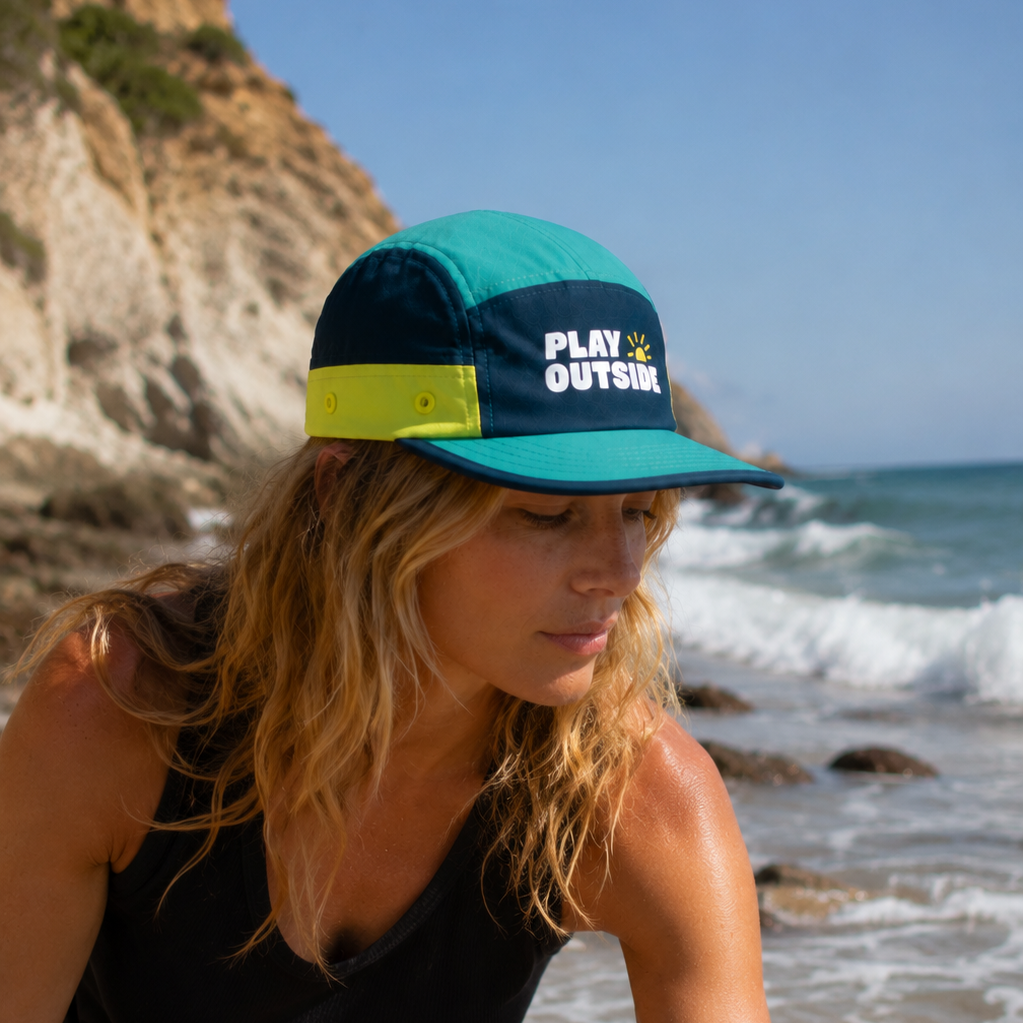 Woman wearing a teal and navy cap with 'Play Outside' logo on a beach.